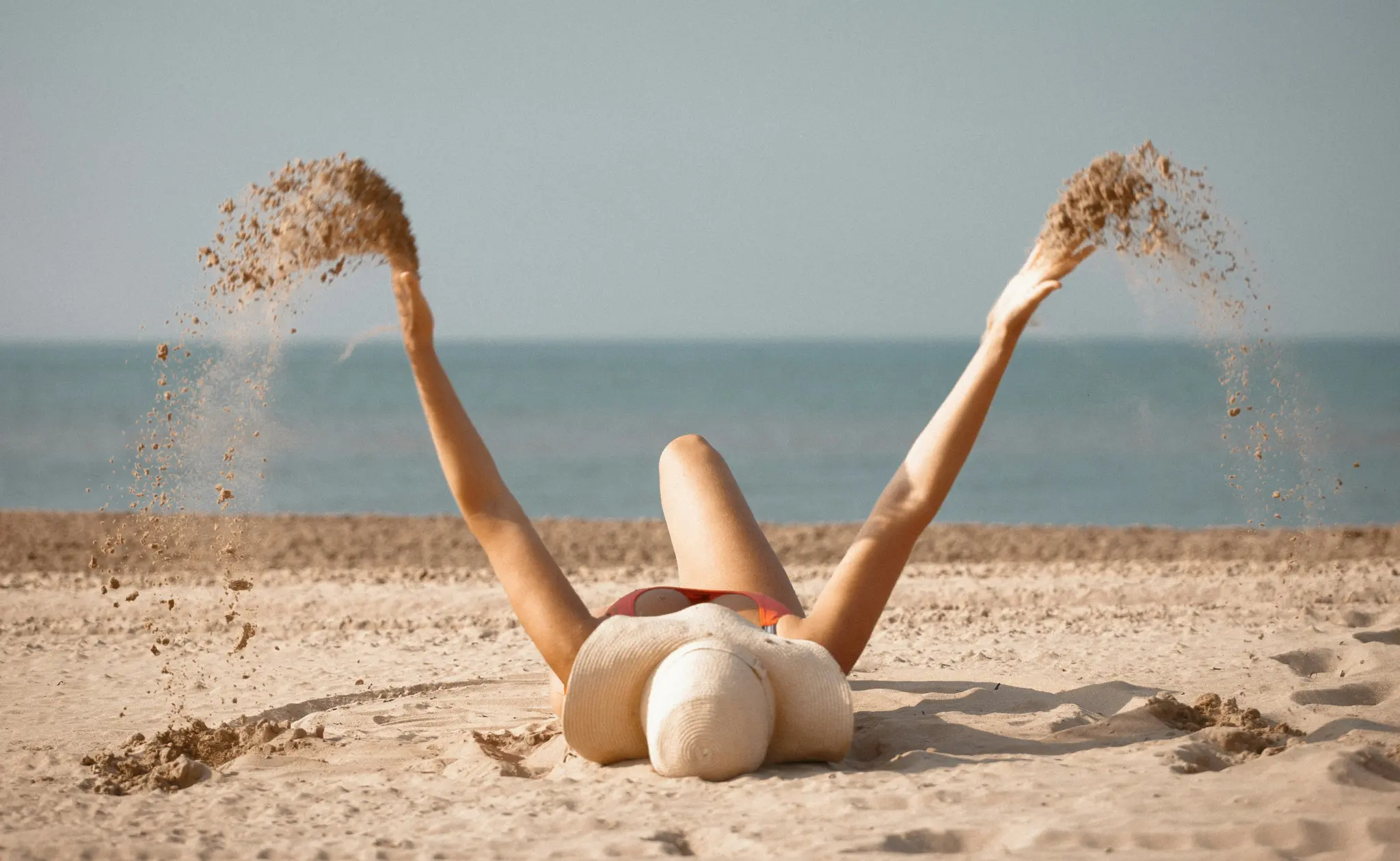 Una persona giace su una spiaggia sabbiosa, indossa un cappello di paglia e un costume da bagno rosso, con le braccia alzate
