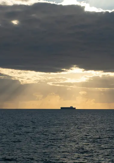 Una grande nave cargo è una silhouette scura all'orizzonte di un oceano blu scuro, sotto un cielo con nuvole drammatiche.