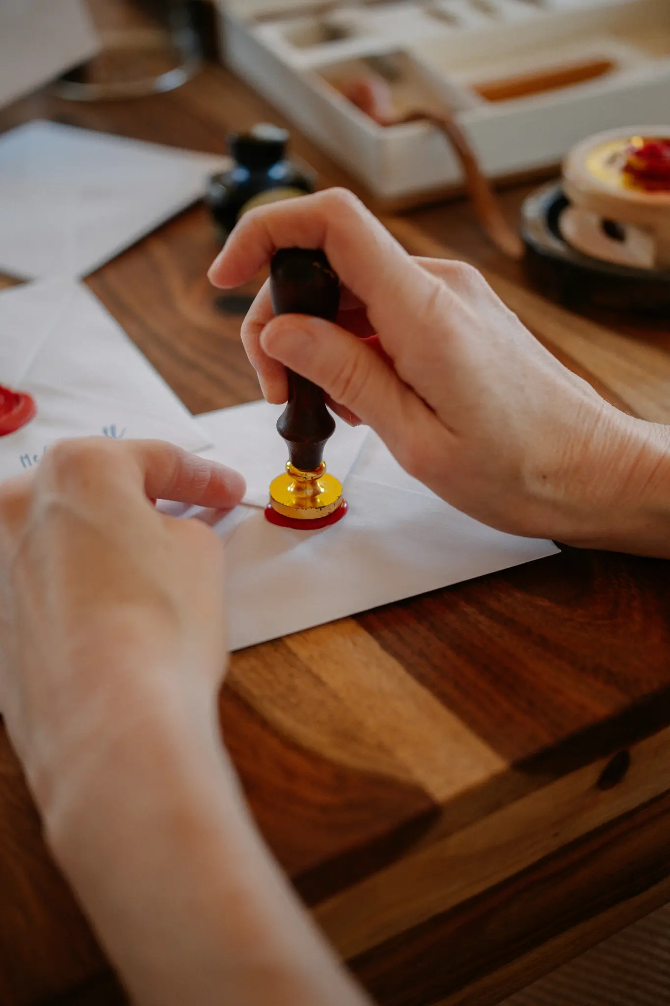 Close-up of hands pressing a gold and dark wood wax seal into red wax on a white envelope on a wooden table.