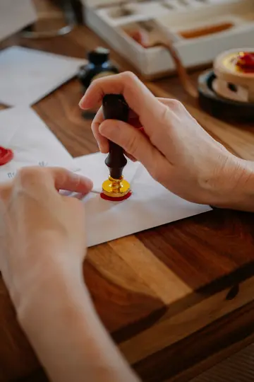 Close-up of hands pressing a gold and dark wood wax seal into red wax on a white envelope on a wooden table.