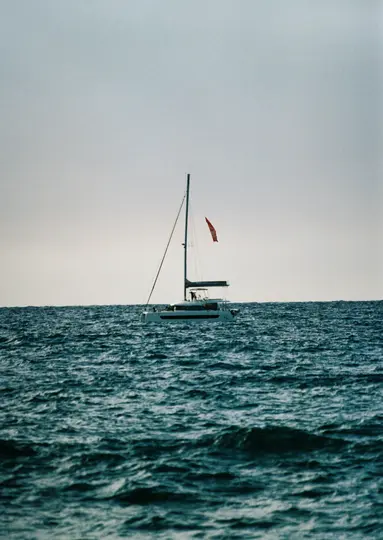 Un catamarano bianco con un albero alto e una piccola bandiera rossa naviga su acqua blu scuro e agitata sotto un cielo