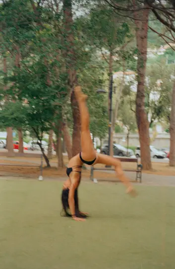 Une photo floue d'une personne en bikini noir faisant un salto arrière sur une surface verte, entourée d'arbres.