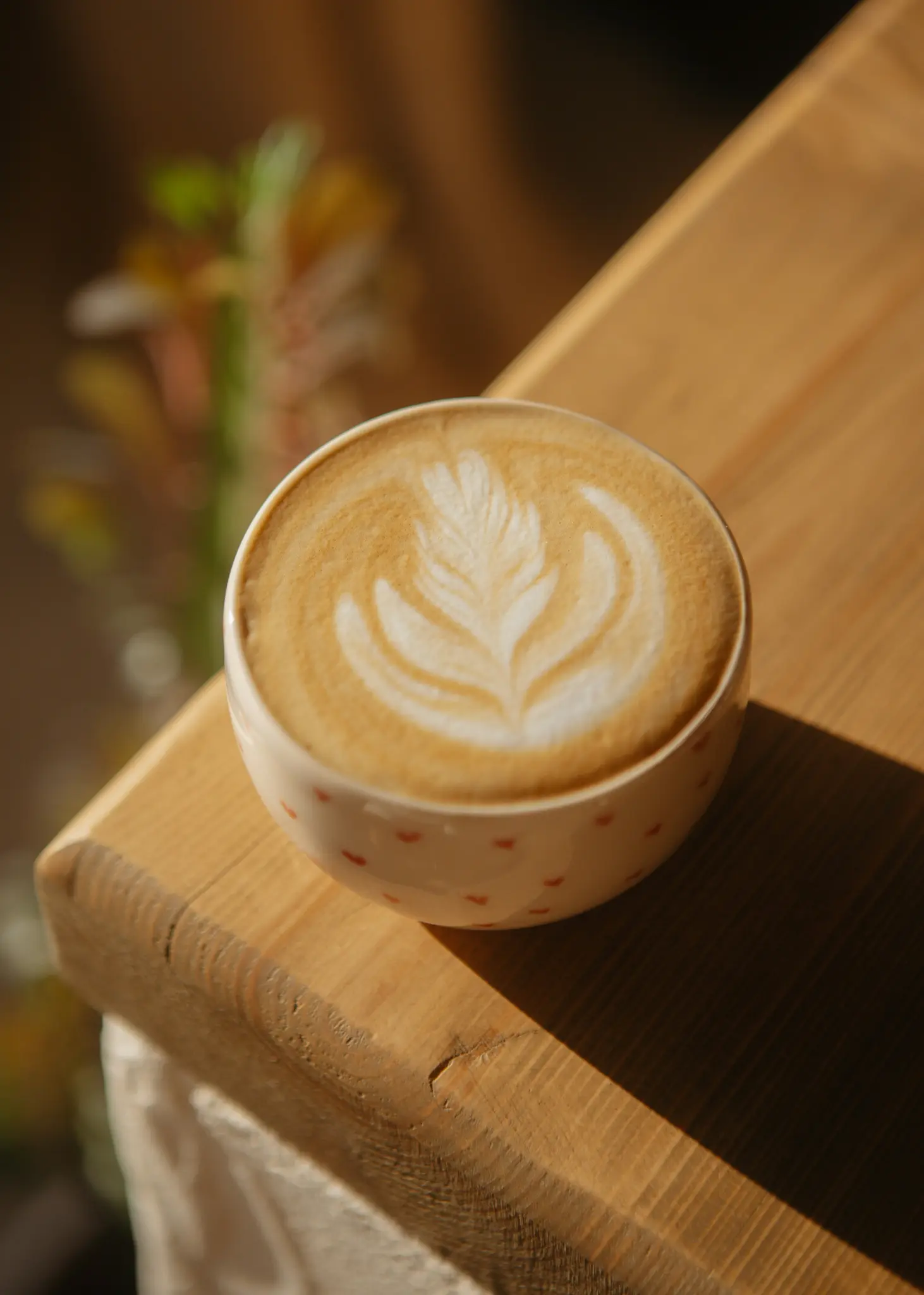 En närbild av en vit keramisk mugg med små röda hjärtan, fylld med latte och intrikat bladformad lattekonst.