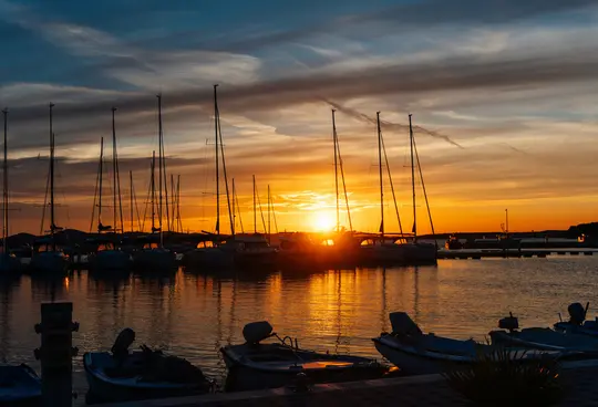 Ein lebendiger Sonnenuntergang über einem Yachthafen, der einen orangefarbenen Schein auf das Wasser wirft und zahlreiche
