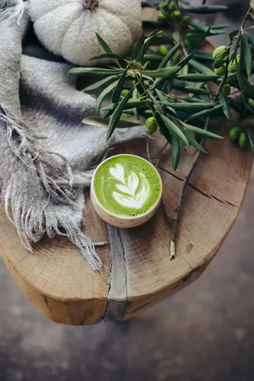 A vibrant green matcha latte with white latte art sits on a rustic wooden surface, surrounded by a gray fringed blanket, a…
