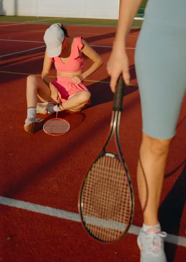 Una persona in rosa è seduta su un campo da tennis rosso, mentre sistema i vestiti. Un'altra persona è in piedi nelle