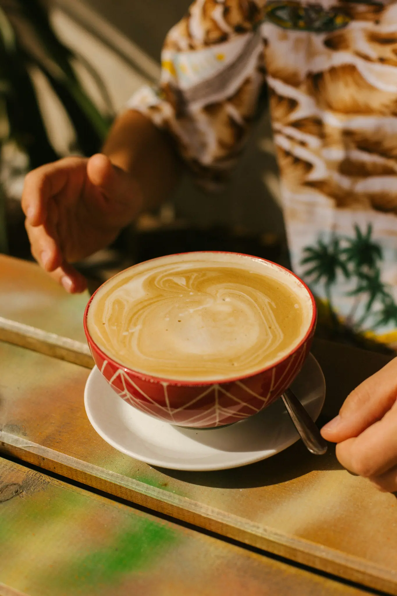 Nahaufnahme einer roten Schale mit Latte Art Kaffee, die auf einer weißen Untertasse auf einem bunten Holztisch steht.