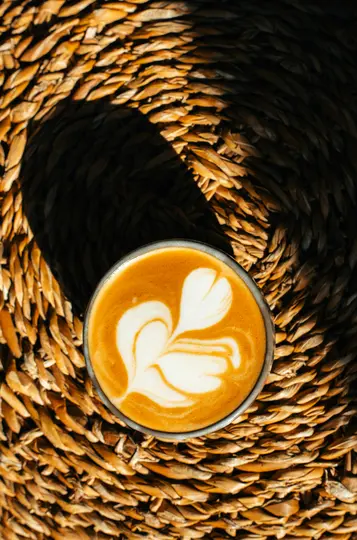 An overhead close-up shot of a latte with white foam art, resembling a heart or leaf, sitting on a textured woven mat.…