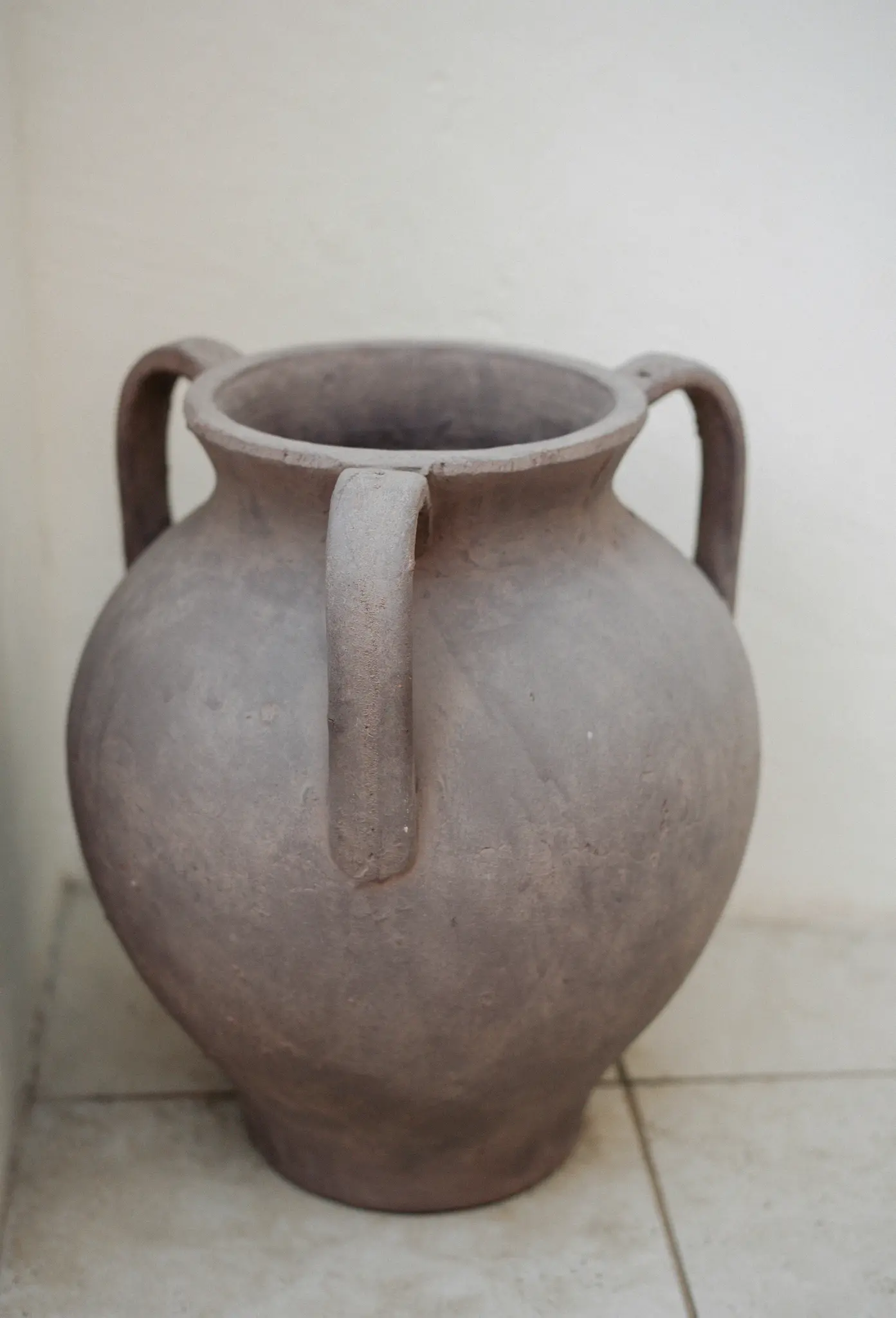 Een grote, ronde, grijze keramische pot met twee handvatten staat op een licht beige betegelde vloer tegen een effen gebroken