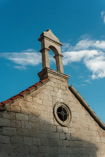 A low-angle shot of an old stone building with a small bell tower and a circular window, set against a bright blue sky with…