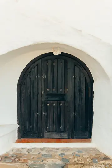Una porta in legno scuro ad arco con accenti metallici è incastonata in un muro di stucco bianco testurizzato. Un piccolo