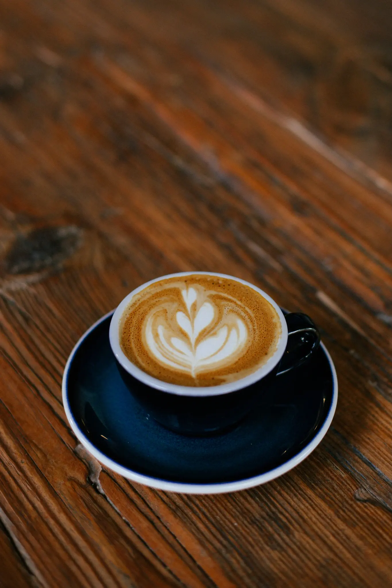 En mørkeblå kop og underkop med en latte med hjerteformet skumkunstdesign, der står på et rustikt træbord.