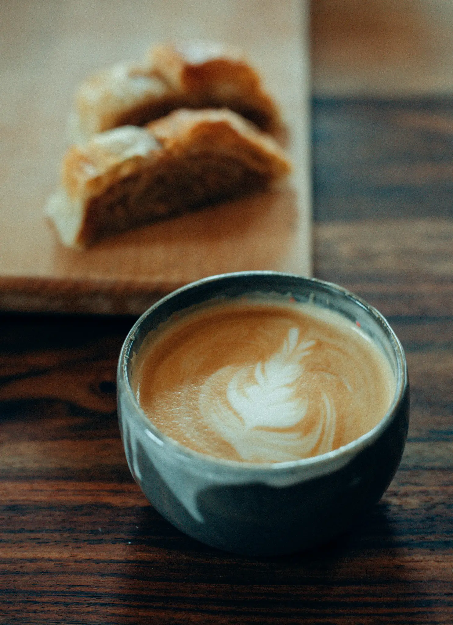 Nahaufnahme einer dunkelgrauen Keramiktasse, gefüllt mit Latte, mit einem zarten weißen Blattmuster auf dem Schaum. Zwei