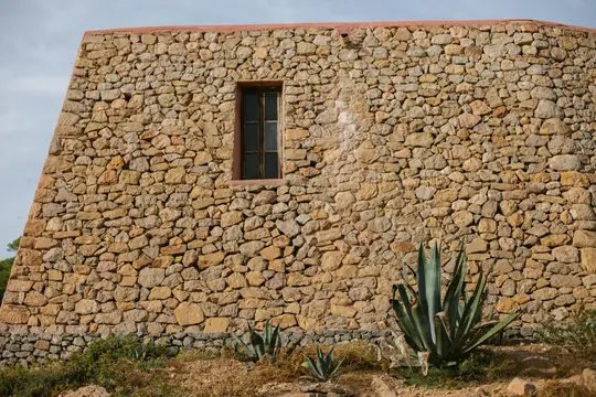 Primo piano di un edificio in pietra con un'unica finestra scura. Diverse piante di agave crescono in primo piano, arido e