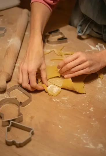 Close-up of hands on a wooden surface, carefully peeling away excess dough from around a cut-out shape. A rolling pin and…