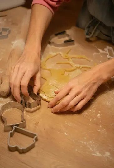 A person's hands are shown from above, pressing metal cookie cutters into light yellow dough on a wooden surface.