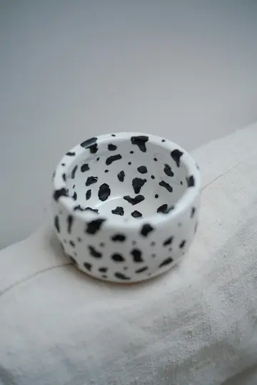 Pieni, pyöreä valkoinen keraaminen kulho, jossa on epäsäännöllisiä mustia pilkkuja, jotka muistuttavat dalmatialaiskuviota