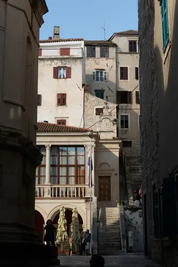 A vertical shot of a narrow street in a historic town. Old buildings with red-shuttered windows line the street, with a…