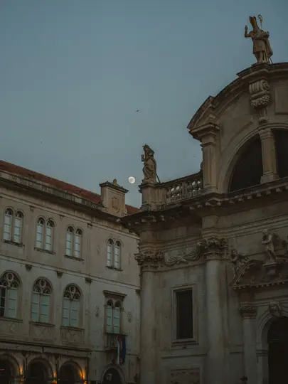 A full moon hangs in a muted blue twilight sky between two ornate stone buildings with statues on their rooftops.