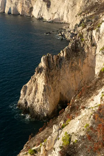 Una vista aerea di imponenti scogliere chiare che si tuffano in un mare blu intenso. Una vegetazione rada e secca si aggrappa