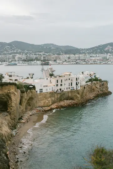 Μια υπερυψωμένη θέα μιας παραθαλάσσιας πόλης με λευκά κτίρια σκαρφαλωμένα σε έναν τραχύ βράχο με θέα έναν ήρεμο κόλπο.