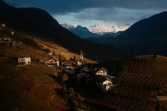 Eine Luftaufnahme eines Dorfes mit Kirchturm, umgeben von terrassierten Weinbergen, in einem Tal mit schneebedeckten Bergen.