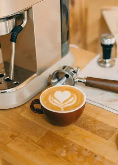 A rich brown ceramic cup holds a latte with intricate white heart-shaped latte art, placed on a light wooden countertop.