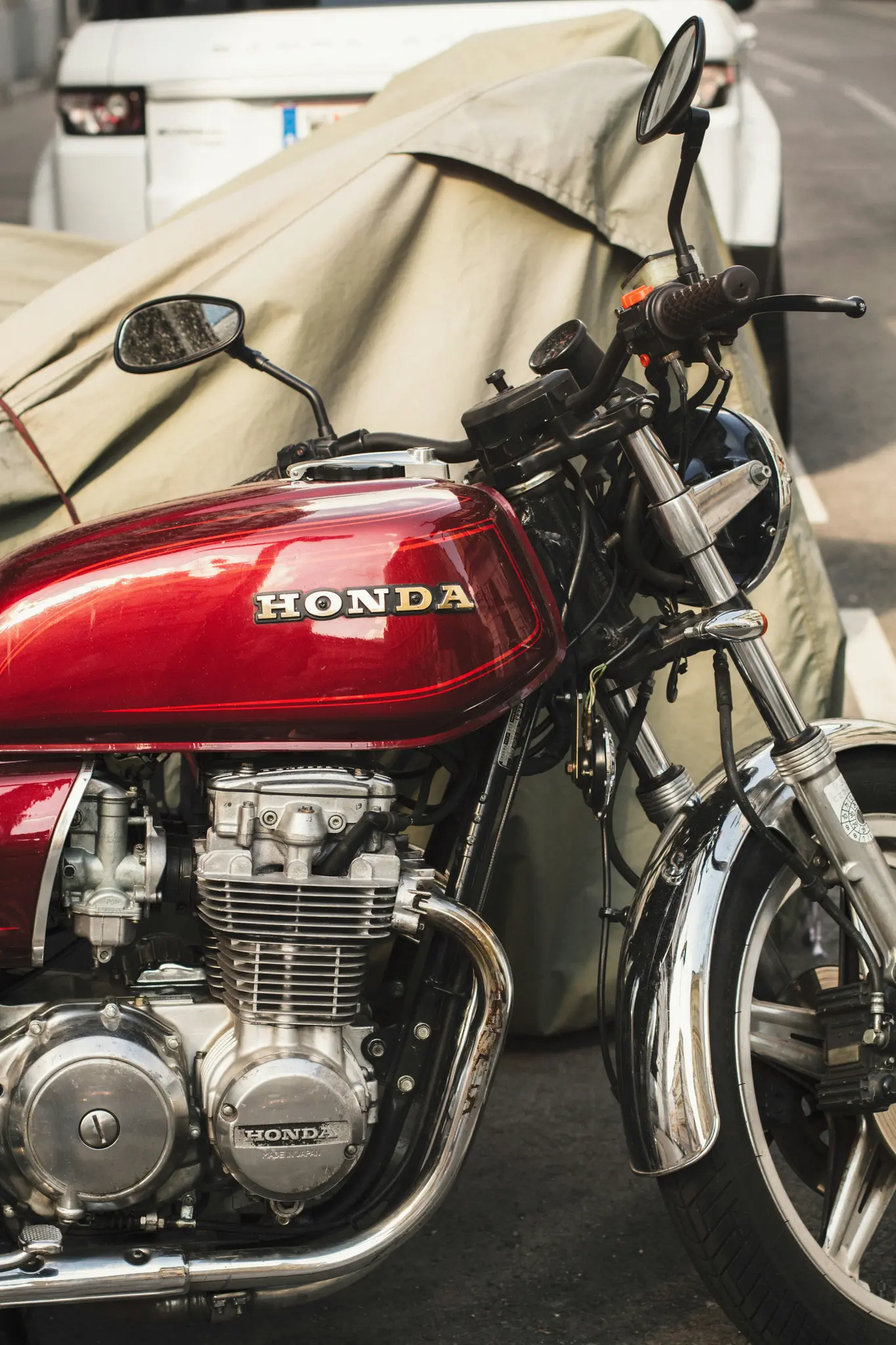Een gedetailleerde close-up van een rode Honda motorfiets met chromen accenten, geparkeerd op straat naast een afgedekte