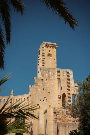Una torre de piedra alta y de color marrón claro con almenas y una fachada desgastada se alza contra un cielo azul claro y