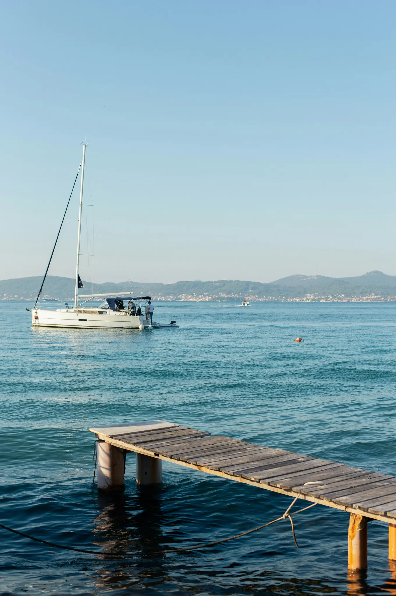 Een witte zeilboot drijft op kalm blauw water met een kleine houten pier op de voorgrond. Verre groene heuvels vormen de