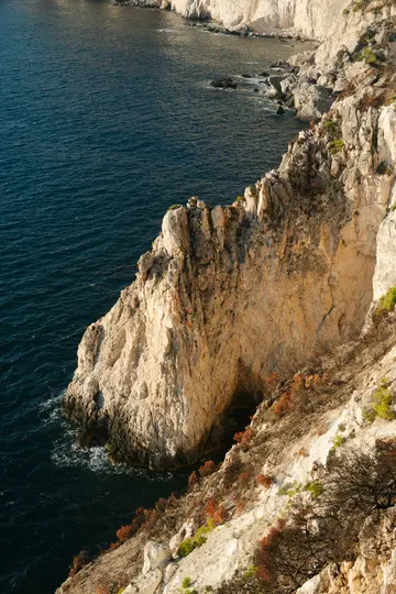 Μια φωτογραφία από ψηλά αποτυπώνει μια δραματική ακτογραμμή με απότομους, ανοιχτόχρωμους βράχους που βυθίζονται σε σκούρο