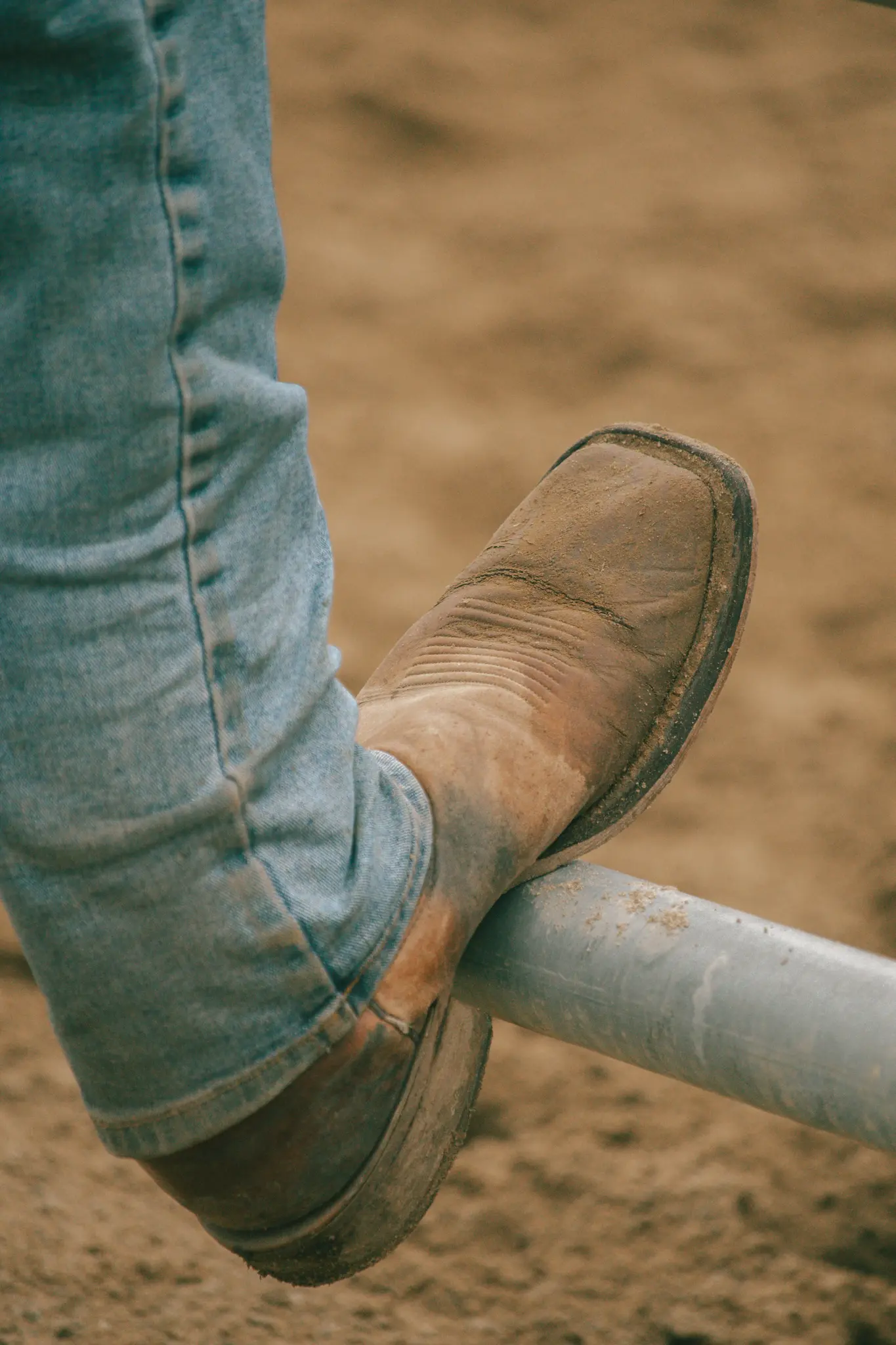 Un primo piano della gamba di una persona, vestita con jeans azzurri e uno stivale da cowboy marrone polveroso, appoggiata su