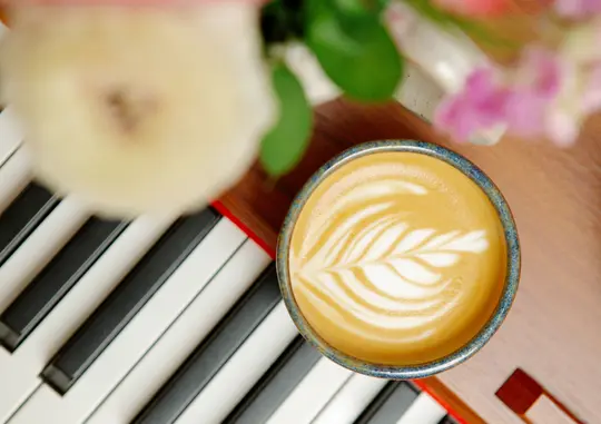 En översiktsbild av en latte med ett lövmönster i skummet, stående på en träyta bredvid svarta och vita pianotangenter.