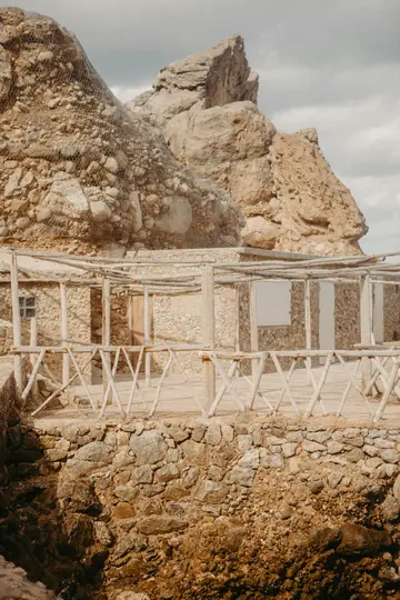 Un edificio in pietra con ringhiera in legno e una parete rocciosa coperta di rete sotto un cielo coperto.