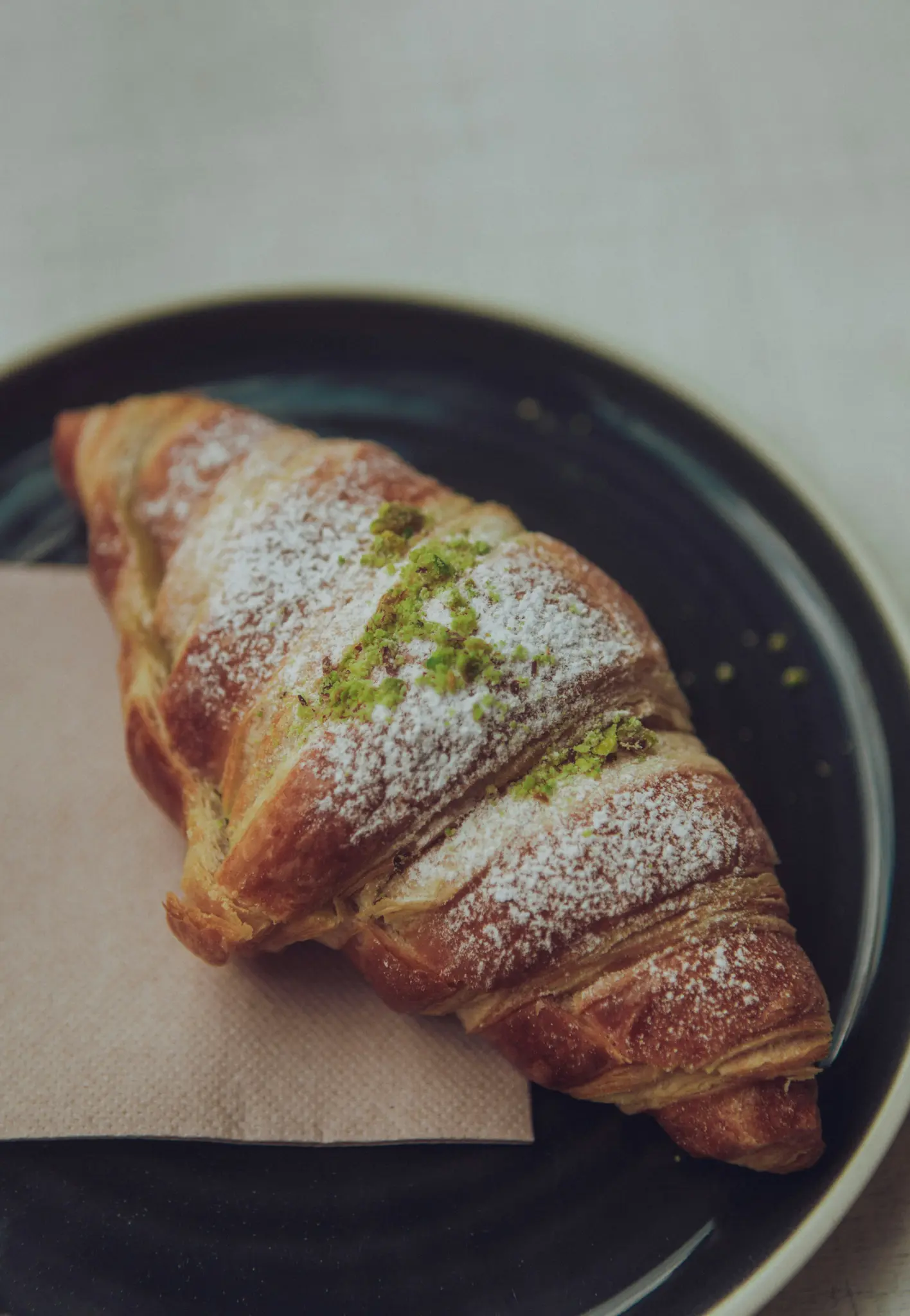 Ein goldbraunes Croissant, großzügig bestäubt mit weißem Puderzucker und leuchtend grünen Streuseln, liegt auf einer beigen