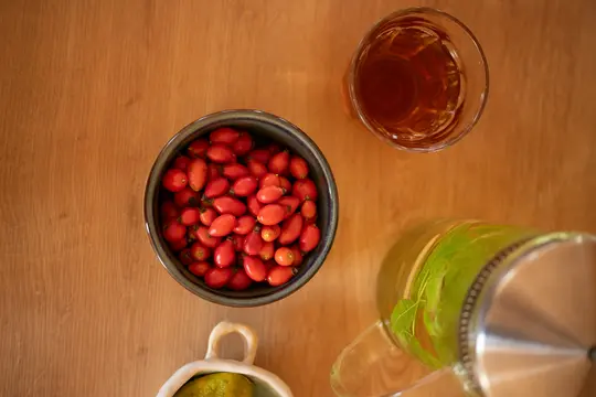 An overhead view of a dark bowl filled with bright red rose hips, a clear glass of amber-colored liquid, and a glass…