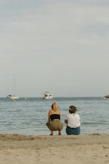 Due donne siedono su una spiaggia sabbiosa di spalle allo spettatore, contemplando il calmo mare blu con barche in