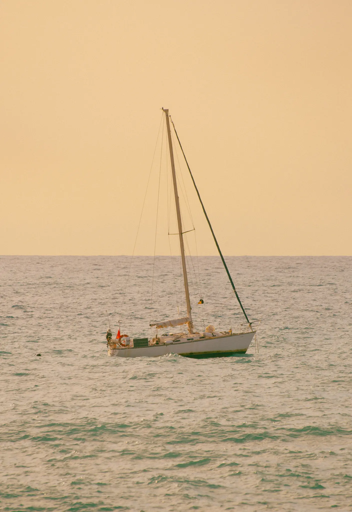 Una barca a vela bianca con un albero alto e vele arrotolate galleggia su acqua turchese leggermente increspata sotto un