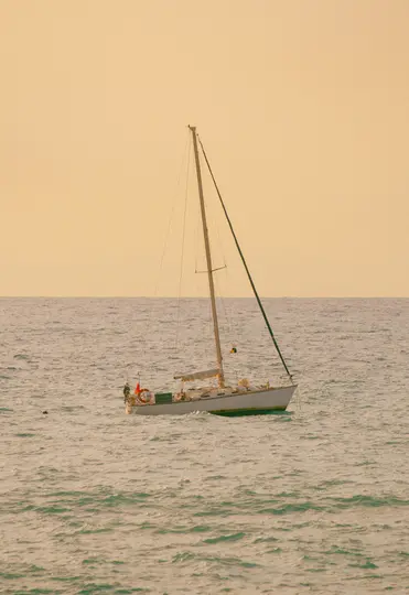 Un velero blanco con un mástil alto y velas enrolladas flota sobre agua turquesa suavemente ondulada bajo un cielo naranja