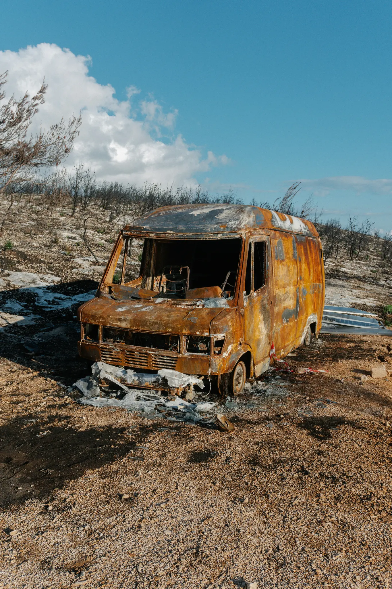 Een zwaar verbrande, roestig oranje en zwarte bestelwagen staat in een dor, door brand verwoest landschap onder een