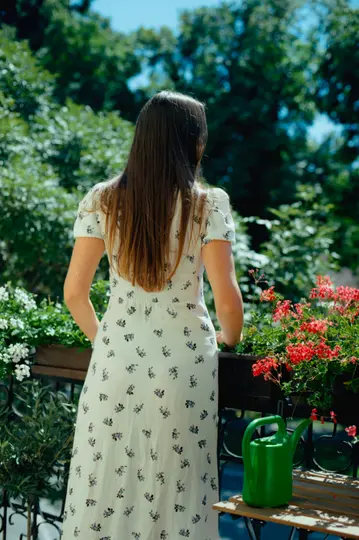 Osoba s dlouhými hnědými vlasy, viděná zezadu, stojí na balkoně a dívá se na zelené stromy. Pokojové rostliny s bílými...