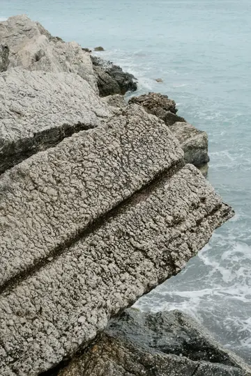Μια κοντινή όψη από ανοιχτόγκριζους, έντονα υφασμένους σχηματισμούς βράχων κατά μήκος της ακτογραμμής, με τη γαλάζια θάλασσα