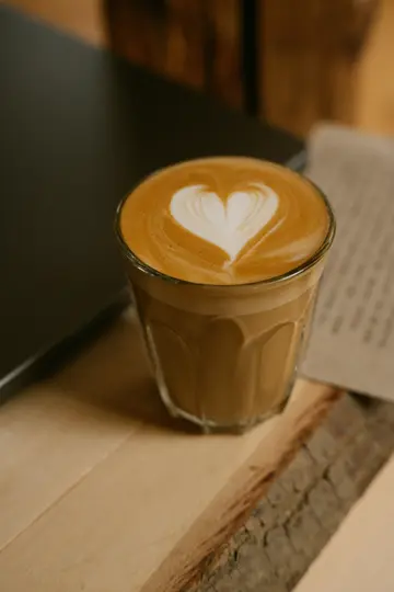 A glass cup filled with a latte, featuring a white heart design in the foam, sits on a light wooden table.