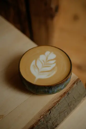 A close-up, slightly overhead shot of a latte in a dark ceramic cup, featuring intricate white leaf latte art on its…