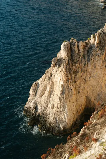 Μια άποψη υψηλής γωνίας ενός τραχύ, ανοιχτόχρωμου βράχου που βυθίζεται σε σκούρο μπλε ωκεάνιο νερό. Είναι ορατή αραιή
