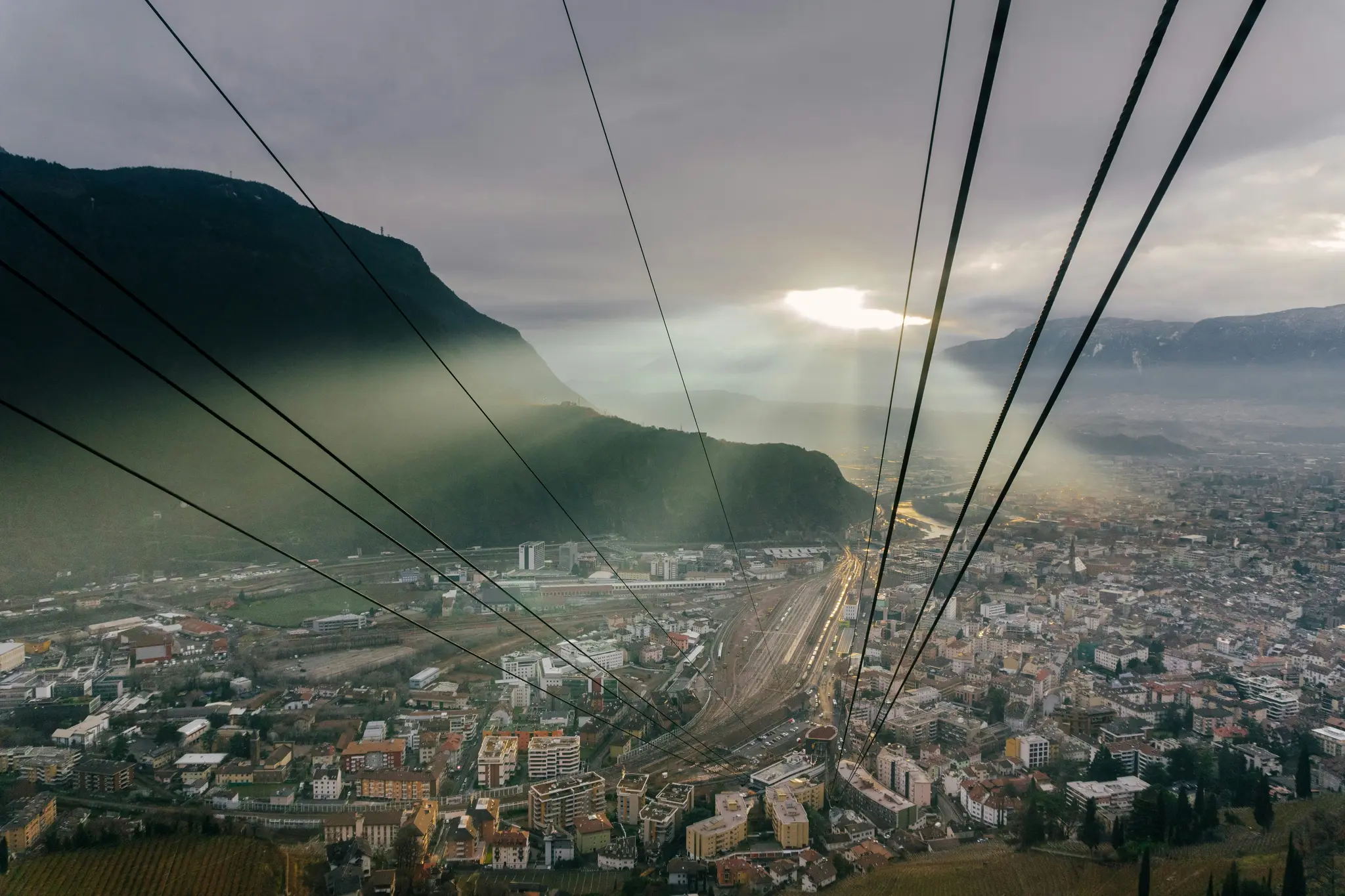 Een luchtfoto van een stad in een vallei, omringd door bergen onder een bewolkte hemel met doorbrekende zonnestralen.