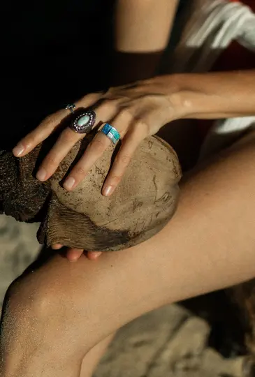 Close-up of a person's hands adorned with rings, holding a coconut. The person's legs are visible in the foreground.