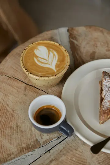 Un primo piano dall'alto di un latte con un disegno a foglia nella schiuma, una tazzina di espresso e una fetta di torta.
