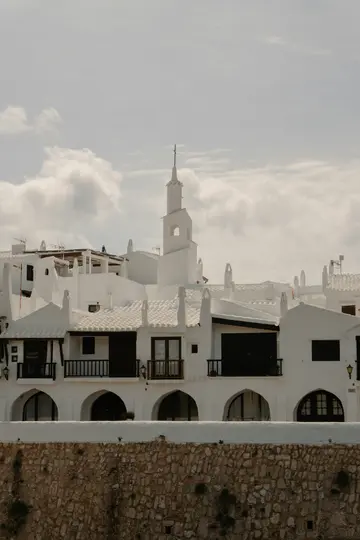 A picturesque village scene featuring numerous white buildings with dark windows and balconies, and a tall bell tower.