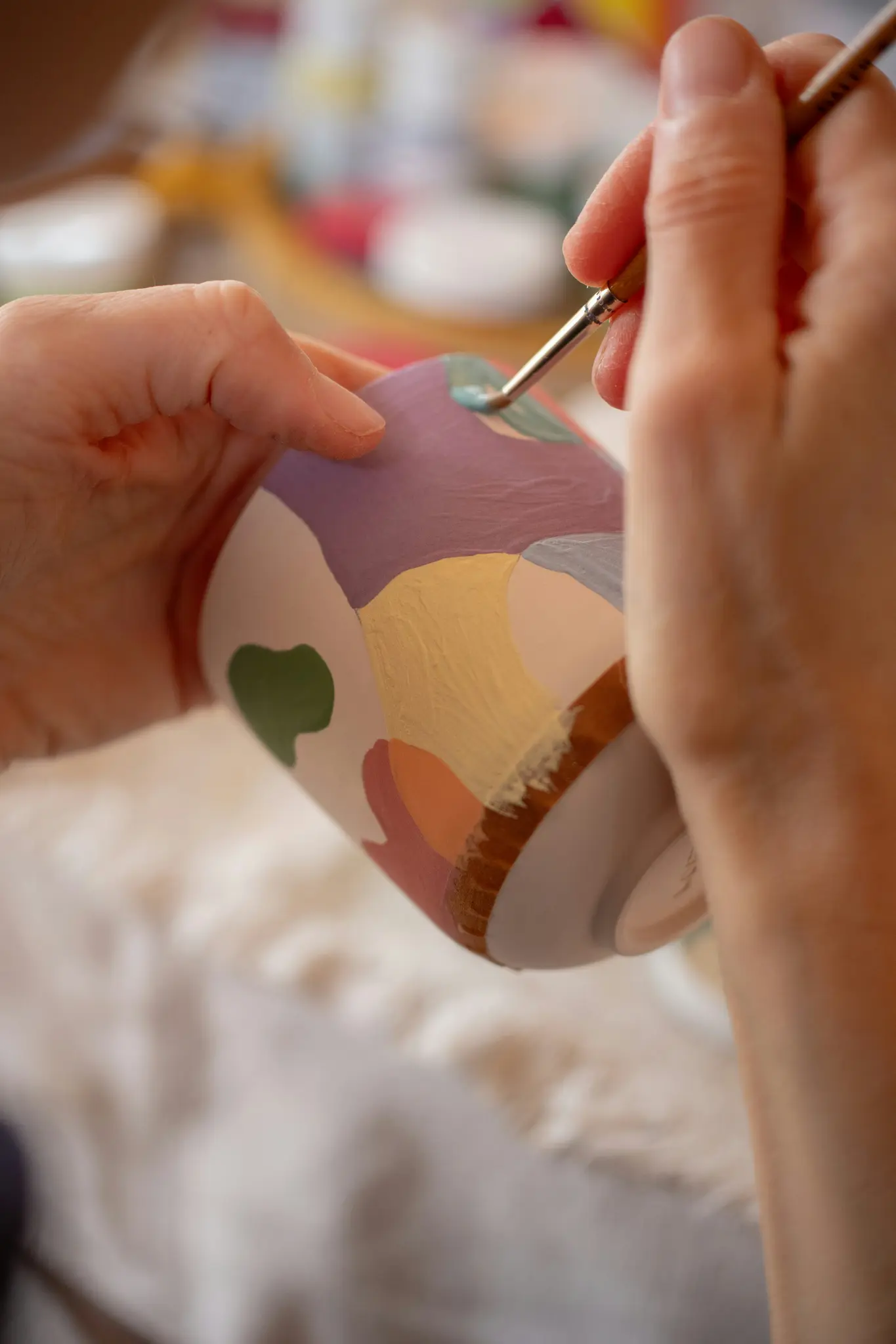 Gros plan sur des mains peignant une tasse en céramique avec des formes abstraites dans des couleurs pastel et terreuses. Une